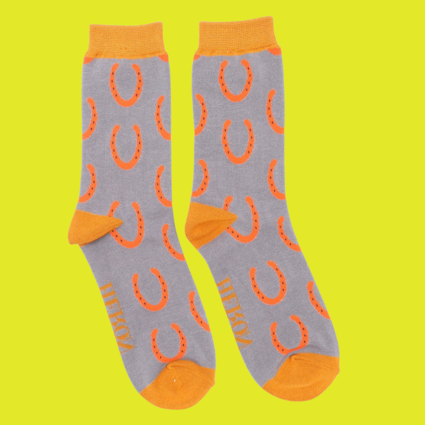 Horseshoes Bamboo Socks - Miss Sparrow. A pair of grey socks featuring an orange horseshoe pattern. Orange cuffs, heels, and toes. Socks shown on a yellow background.