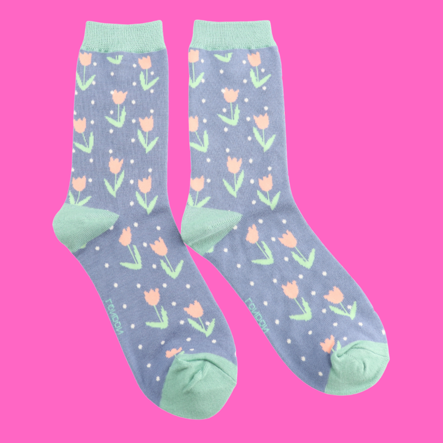 Tulips Bamboo Socks - Miss Sparrow. A pair of light blue socks featuring white polka dots, and a light pink and green tulips pattern. Light green cuffs, heels, and toes. Socks shown on a pink background.