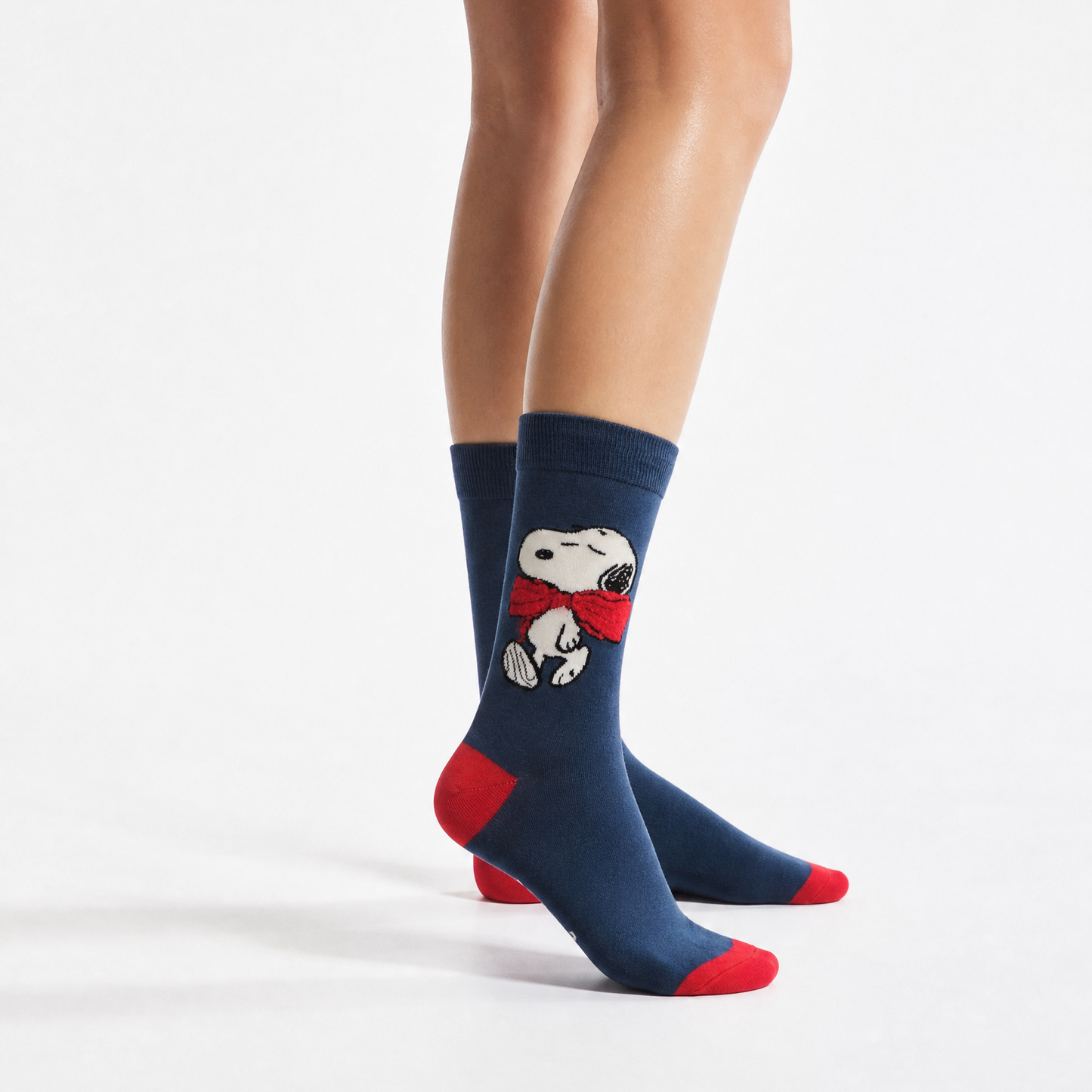 A person's legs wearing navy blue crew socks with red toes and heels, featuring snoopy on the side, against a plain white studio background.