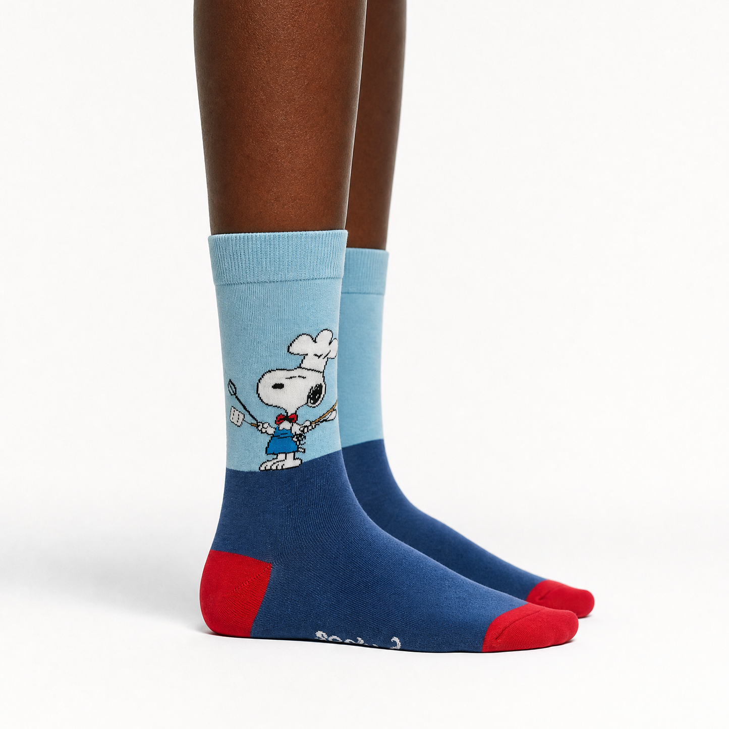 A pair of crew socks worn on a person's feet, shown against a clean white studio background. The socks are light blue at the top with snoopy in a chefs hat, dark blue through the foot, and bright red toes and heels.