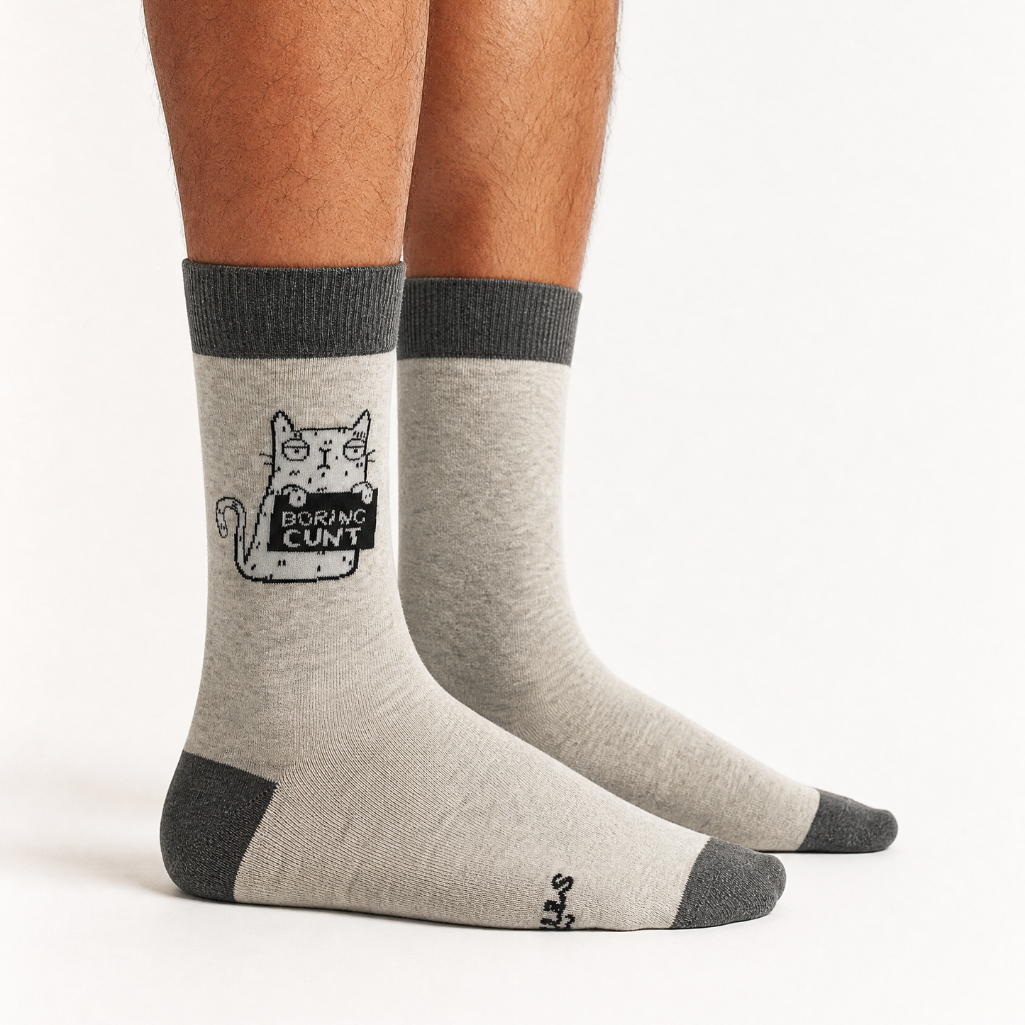 A pair of crew socks worn on a person's feet, shown against a plain light background. The socks are heather light grey with dark grey cuffs, heels, and toes, featuring a black-and-white cartoon cat graphic on one sock that reads 'BORING CUNT'.