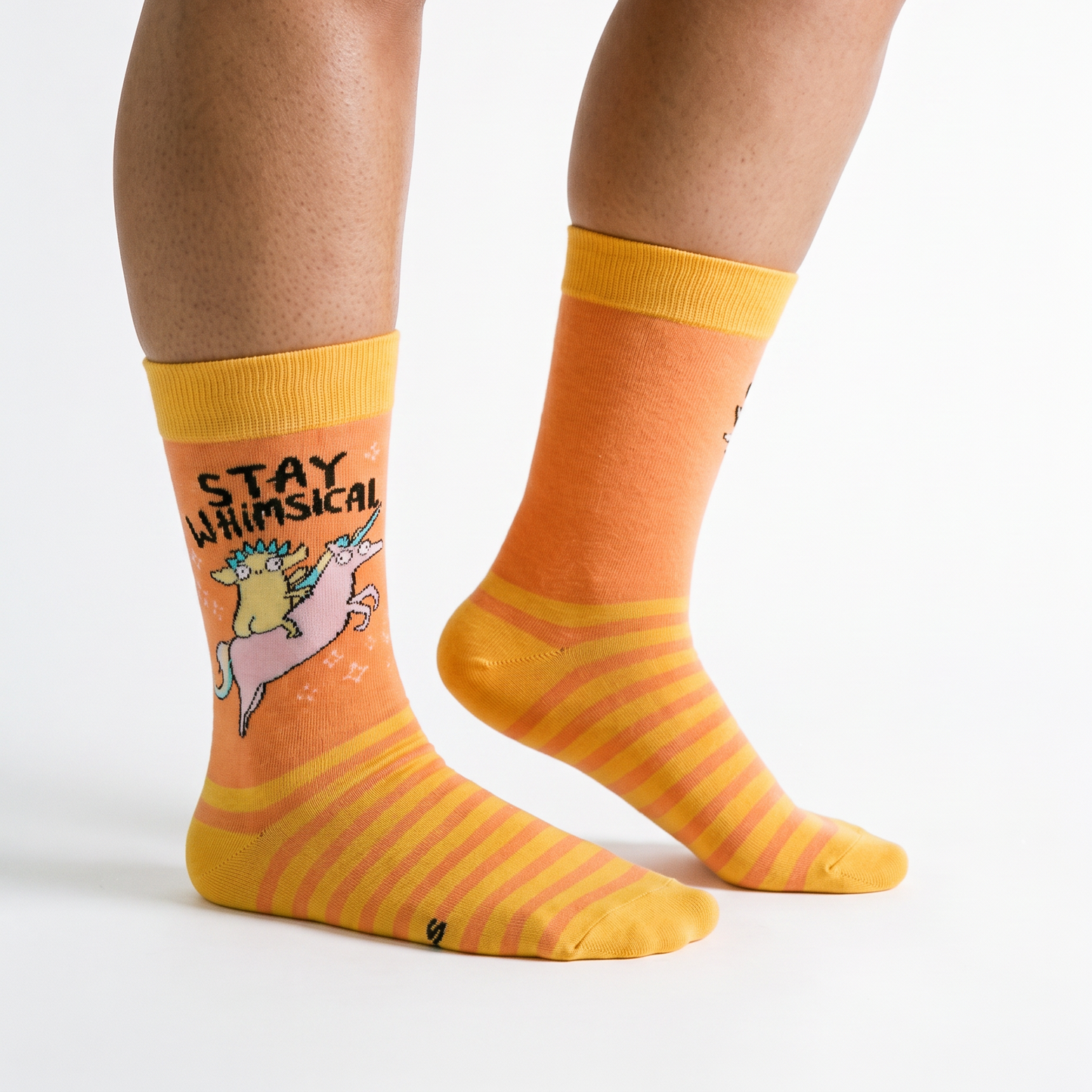 A pair of crew socks worn on a person's feet, shown against a clean white studio background. The socks are predominantly orange and yellow with a graphic on one sock featuring the text 'STAY WHIMSICAL' above a pink unicorn.