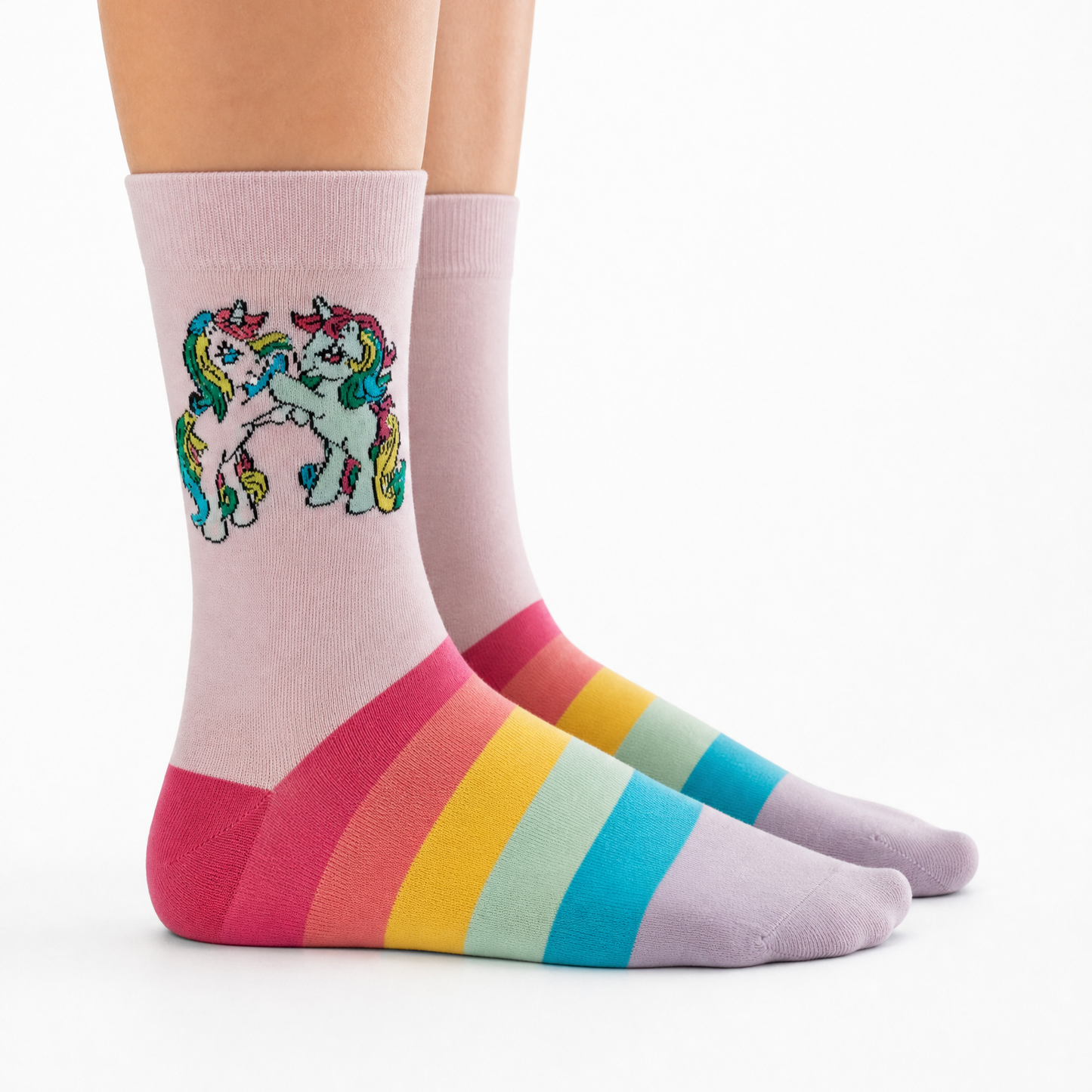 A pair of crew socks worn on feet, featuring a pastel pink base with rainbow stripes at the heel, toe, and sole, and a colorful embroidered unicorn design on the front sock.