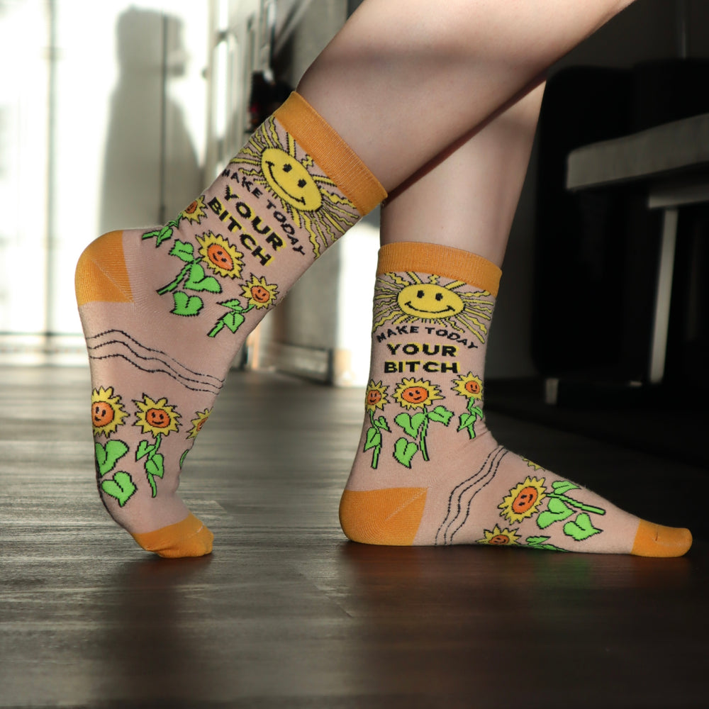 
                      
                        Make Today Your B**tch Socks. Model wearing a pair of socks depicting the sun and sunflowers with a bold sweary slogan. Orange toes, cuff and heel.
                      
                    