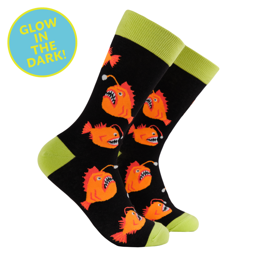 A pair of black crew socks with bright lime green cuffs, heels, and toes, featuring a repeating pattern of orange glow-in-the-dark anglerfish. A blue circular badge in the top left corner reads 'GLOW IN THE DARK!'.