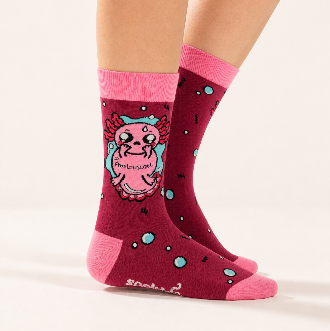 A pair of crew socks worn on a person's feet, shown against a plain light beige studio background. The socks are dark pink with bright pink cuffs and toes, featuring a cartoon creature graphic and light blue bubble accents.
