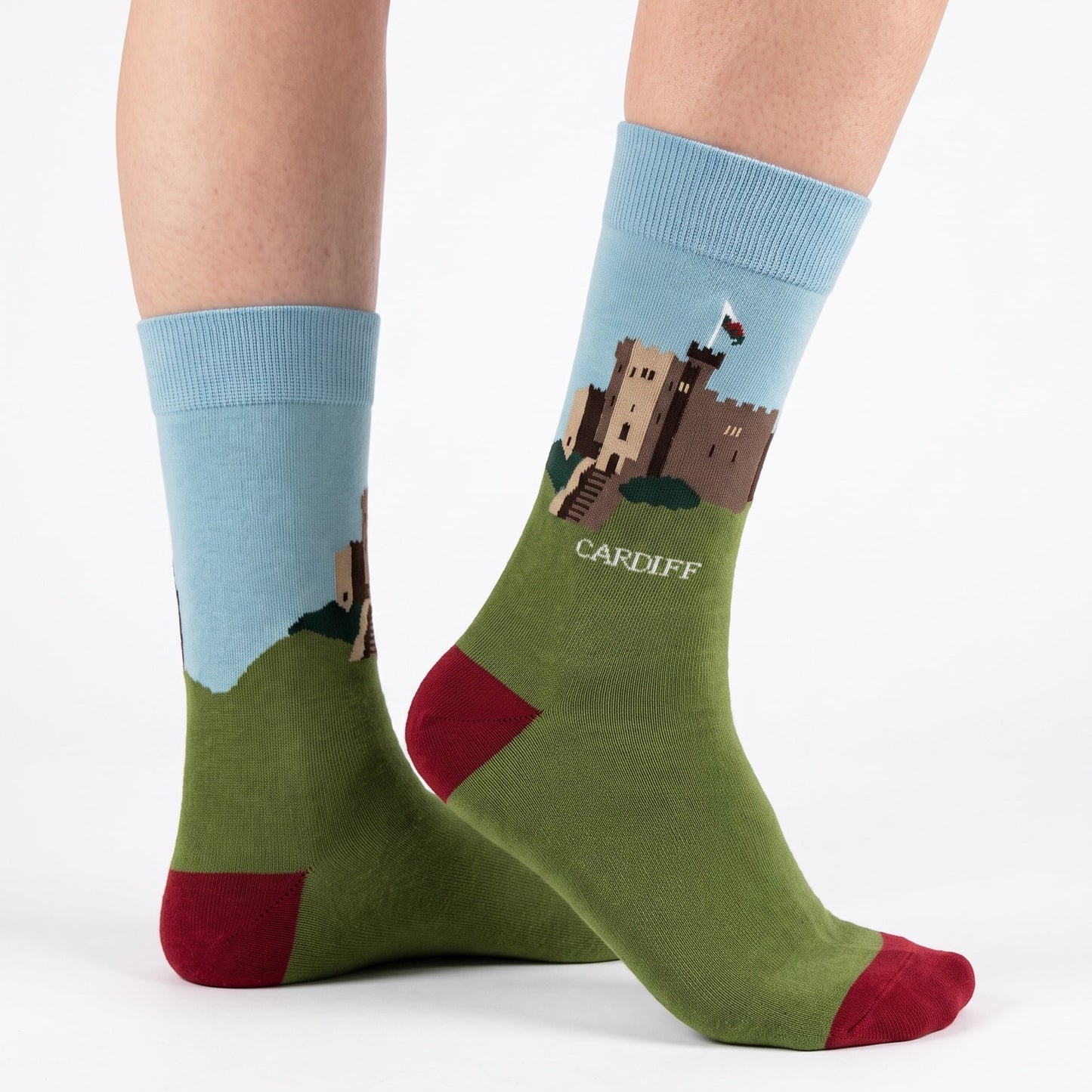 A pair of crew socks worn on a person's feet, featuring a light blue cuff and a green lower section with a printed illustration of Cardiff Castle and the text 'CARDIFF'. The toes and heels are solid red.
