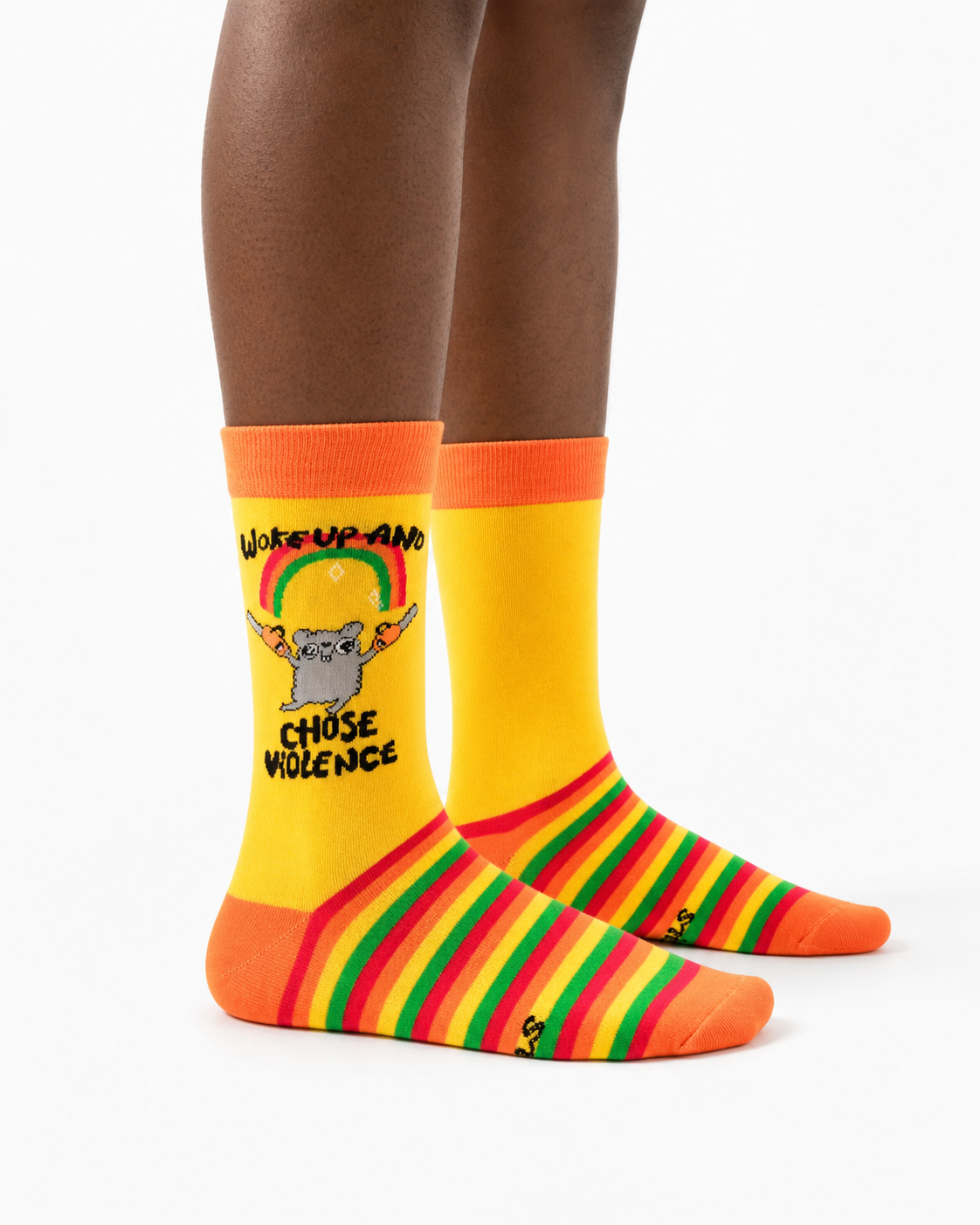 A pair of crew socks worn on a person's feet, shown against a plain white studio background. The socks are bright yellow with orange cuffs and toes, green and red diagonal stripes on the lower half, and a cartoon cat graphic with the text 'Wake Up AND CHOOSE VIOLENCE'.