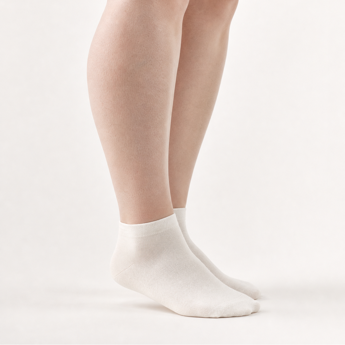 A close-up of a person's legs wearing a pair of plain white ankle socks against a clean, light off-white studio background.