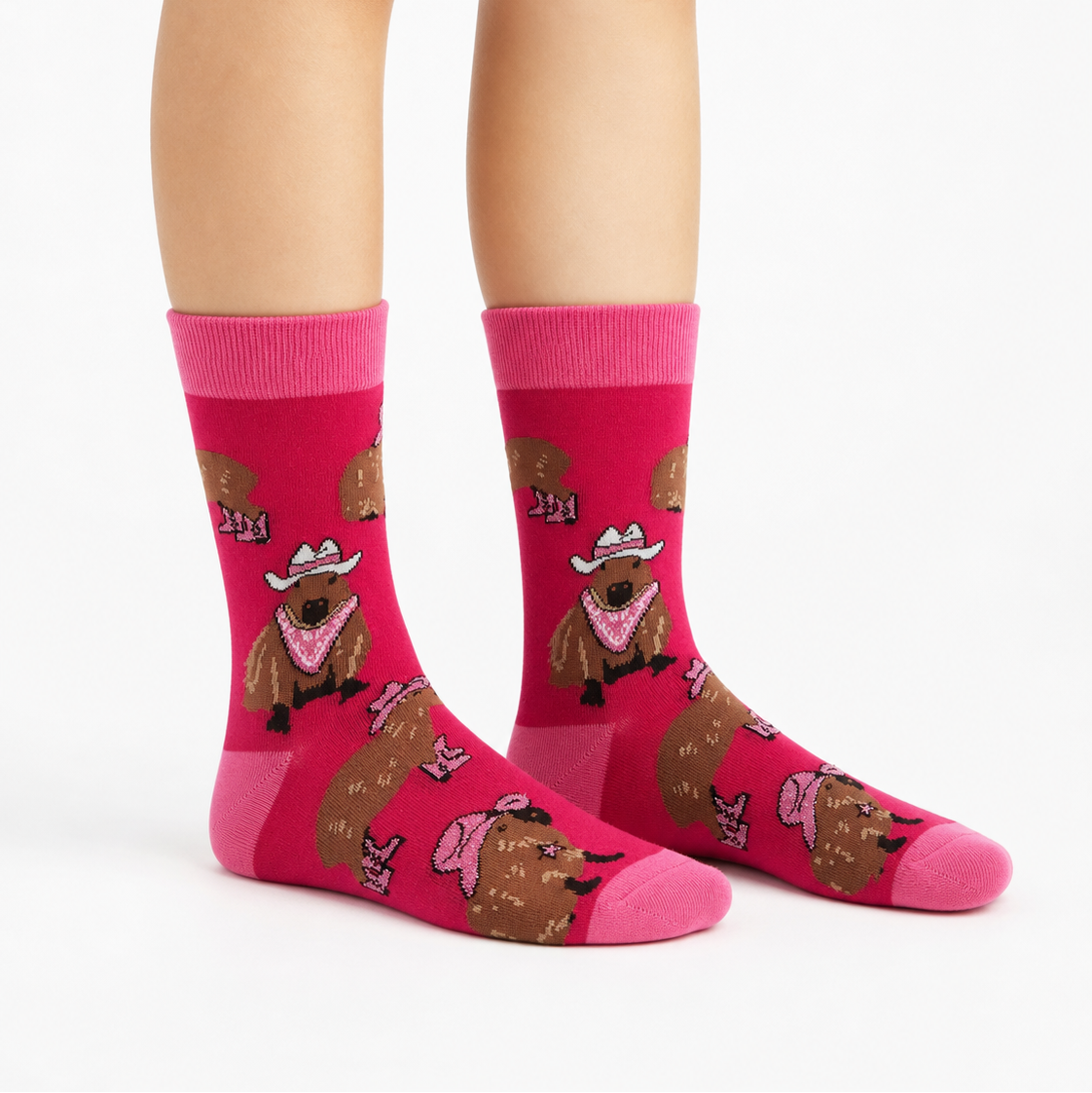 A pair of bright pink crew socks with a repeating cartoon cowboy capybara pattern, shown on a person's feet against a clean white studio background.