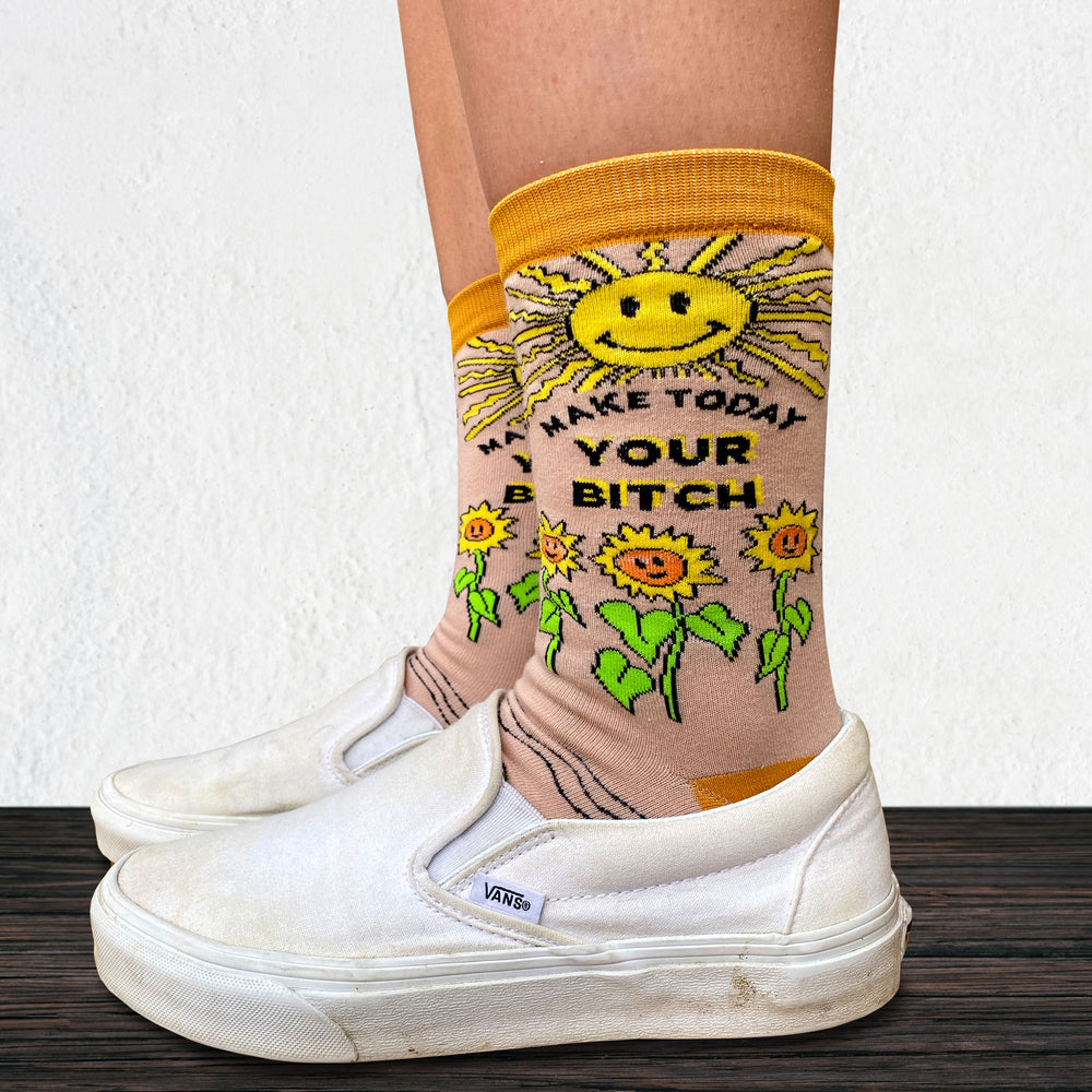 
                      
                        Make Today Your B**tch Socks. Model wearing a pair of socks depicting the sun and sunflowers with a bold sweary slogan. Orange toes, cuff and heel.
                      
                    