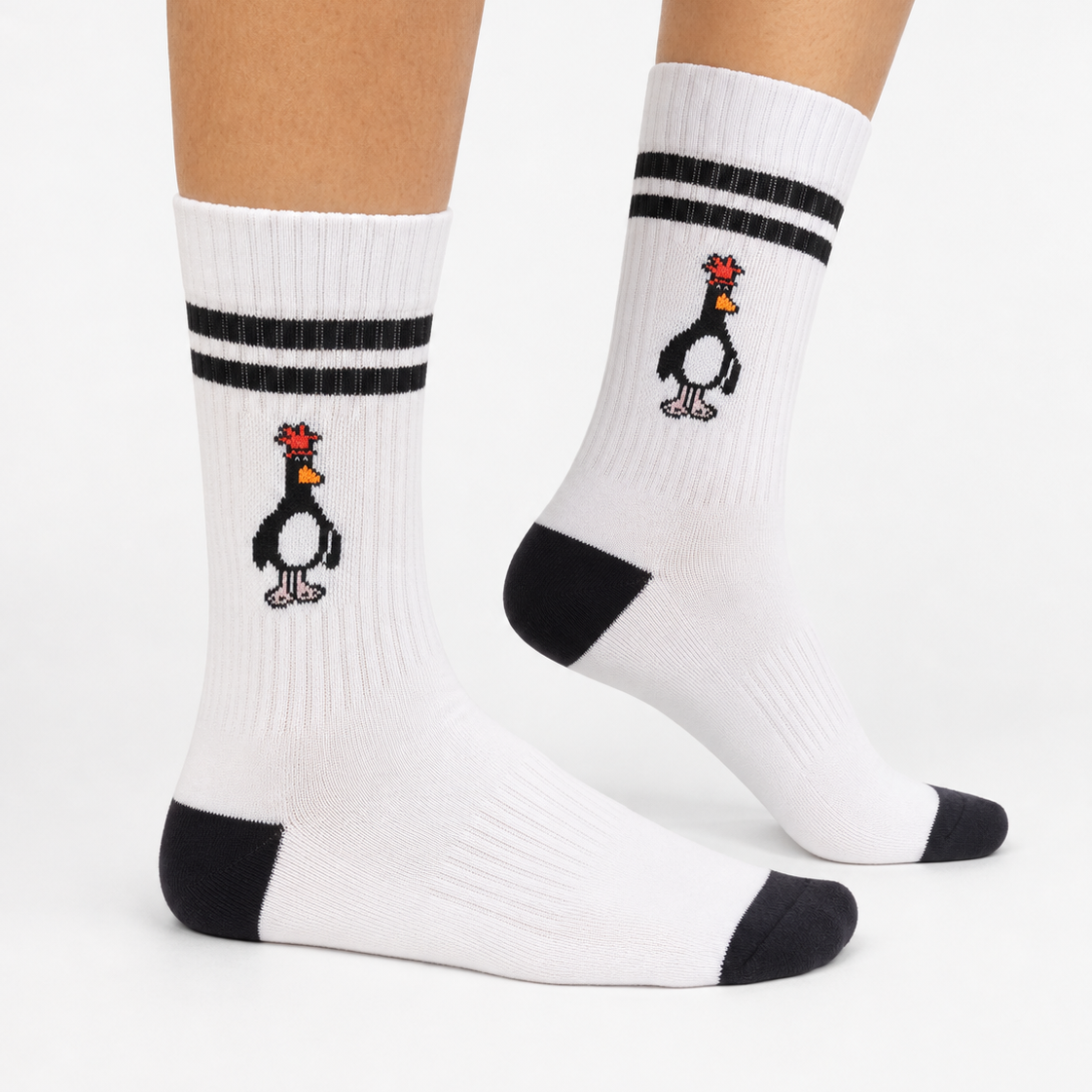 White crew socks with black stripes near the top and a black-and-white penguin graphic with a red comb, shown on a person's feet against a light gray studio background.
