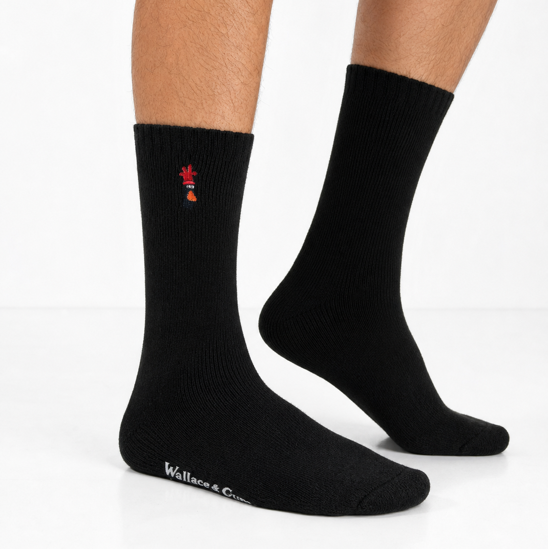 A pair of black crew socks worn on a person's feet, shown against a clean white studio background. One sock has a small red and blue embroidered logo near the cuff, and the other shows white 'Walcott & Girty' branding near the toe.