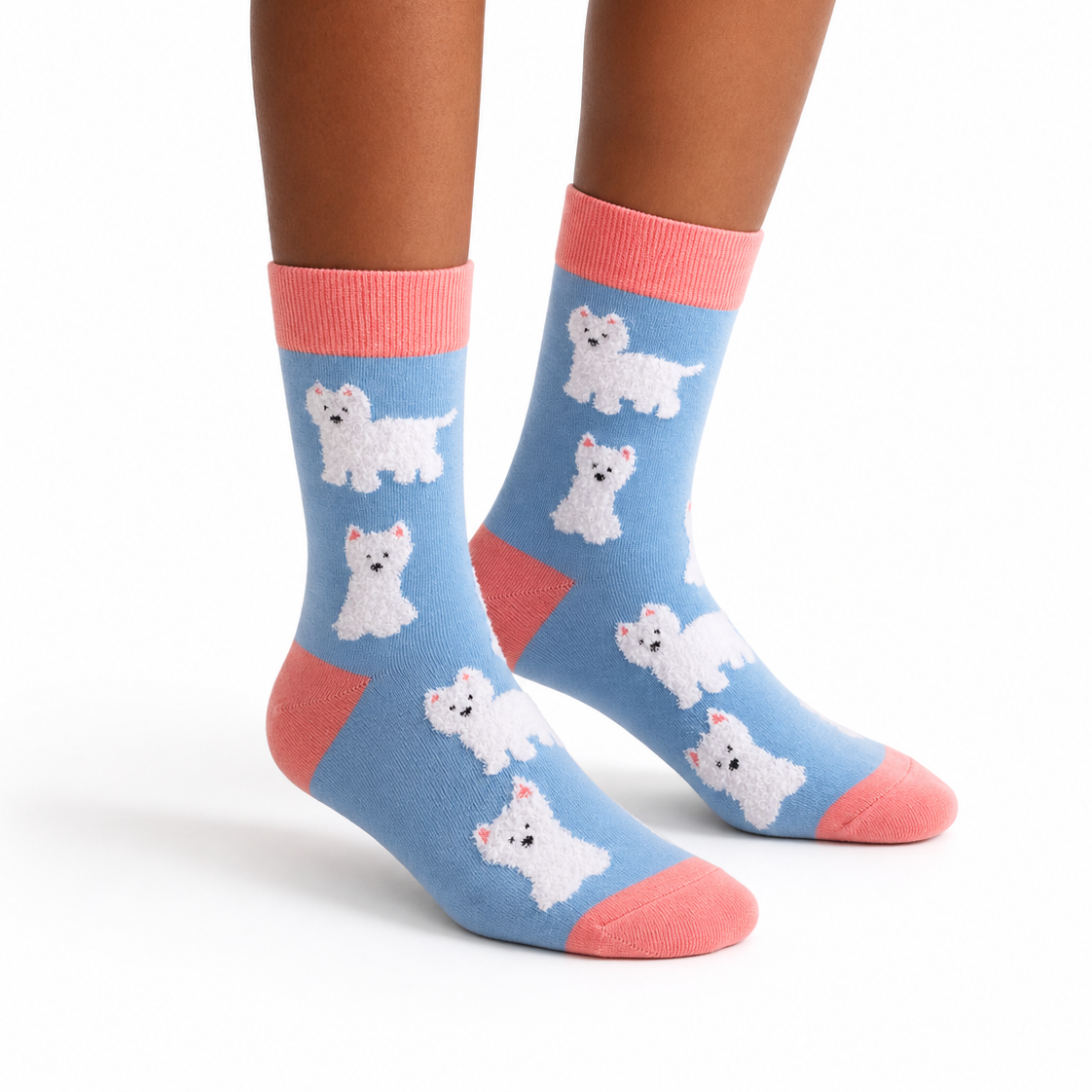 A pair of crew socks worn on a person's feet, featuring a light blue background with white West Highland terrier dog illustrations, coral pink cuffs, heels, and toes, shown against a plain white background.