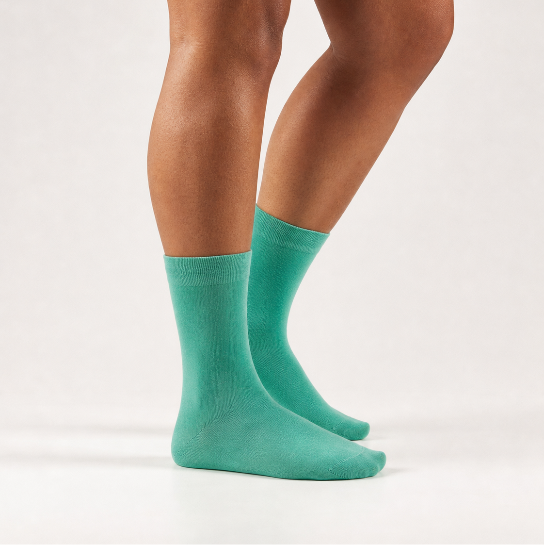 A person's legs wearing a pair of solid teal crew socks, shown from the calves down against a clean, light off-white studio background.