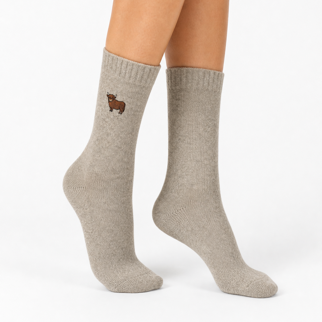 A pair of light taupe crew socks worn on a model's feet, featuring a small embroidered brown animal logo on the upper cuff, against a clean white studio background.