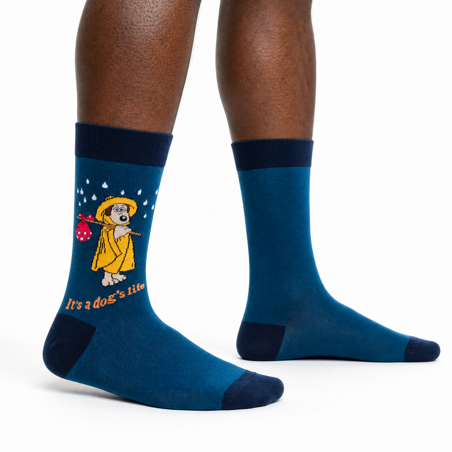A pair of crew socks worn on a person's legs, shown against a plain white studio background. The socks are dark blue with a navy cuff, heel, and toe, featuring a cartoon dog in a yellow raincoat holding a red umbrella with the text 'It's a dog's life'.