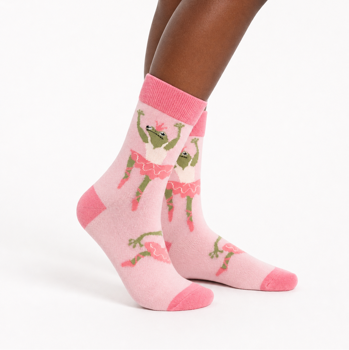 A pair of light pink crew socks with a repeating pattern of green cartoon frogs in pink tutus, worn on a model's feet against a plain white background.