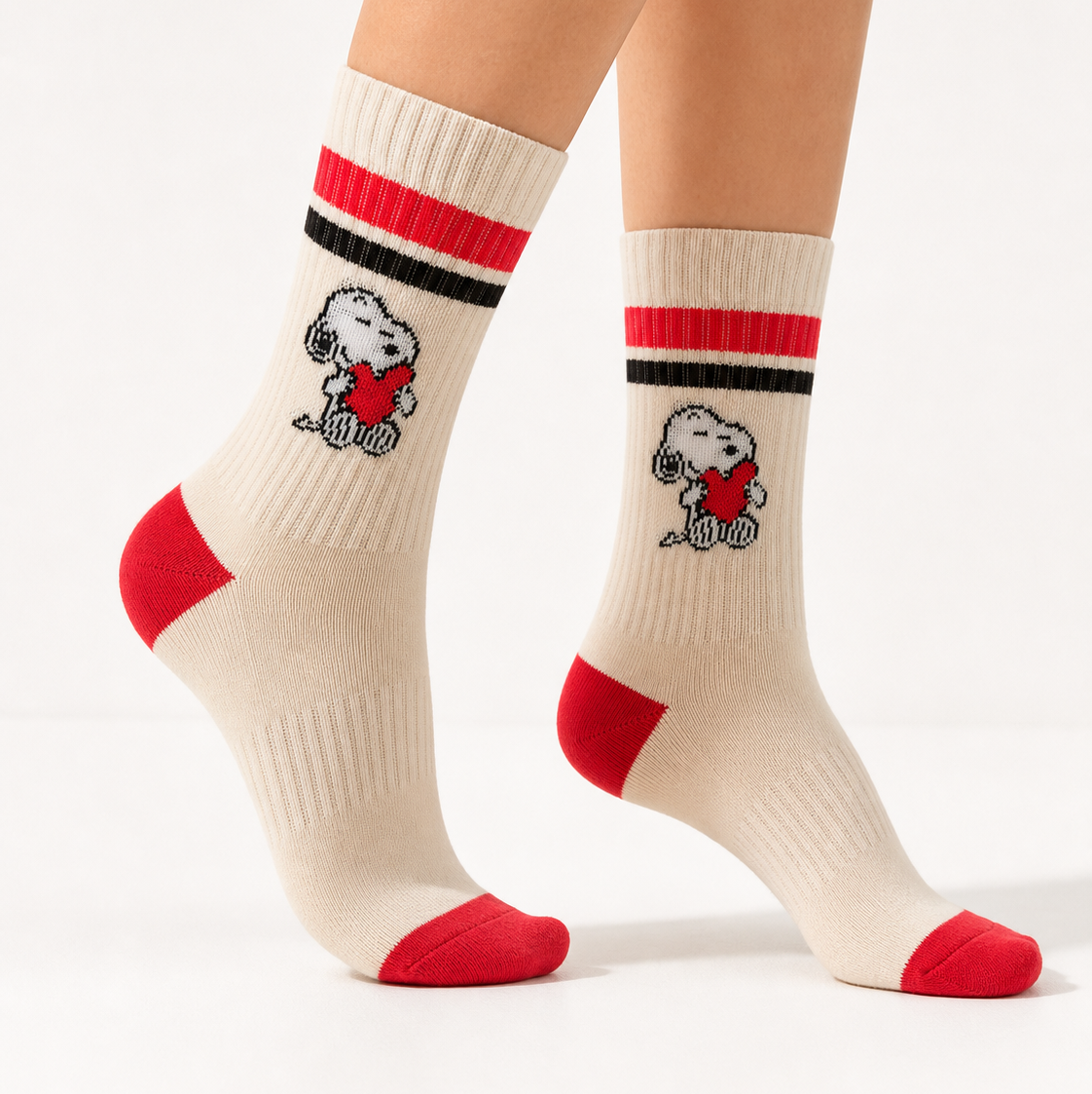 A pair of cream crew socks with red and black stripes near the top, featuring a cartoon dog character wearing a red heart, shown on a model's feet against a white studio background.