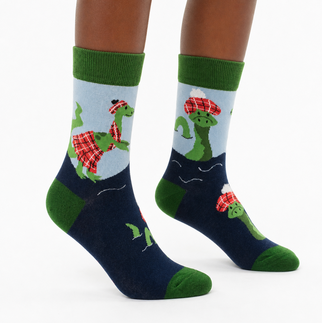 A pair of crew socks worn on a person's feet, featuring a cartoon green dinosaur wearing a red plaid hat and matching plaid scarf against a light blue background, with a dark navy lower section and green heels, toes, and cuffs.