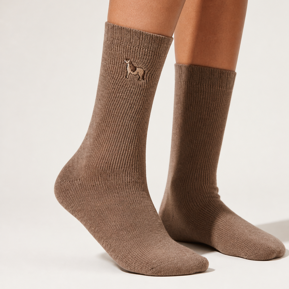 A pair of ribbed crew socks in a warm taupe-brown color, worn on a model's legs. A small embroidered dog logo is visible on the upper cuff of the left sock.