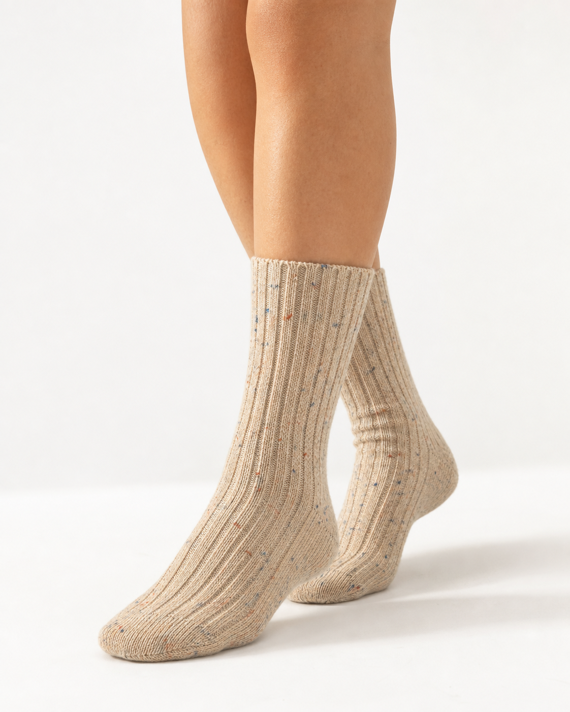 A pair of ribbed ankle socks in a warm beige/cream color with tiny multicolored flecks, worn on a model's feet against a clean white studio background.