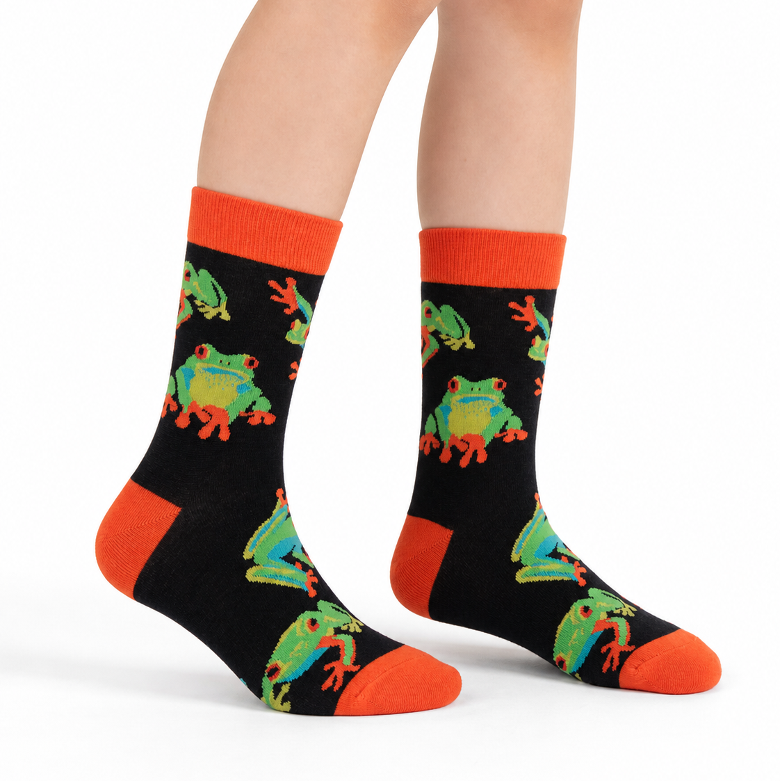 A pair of crew socks worn on a person's feet, featuring a black background with a repeating pattern of green and orange tree frogs, finished with bright orange cuffs, heels, and toes.