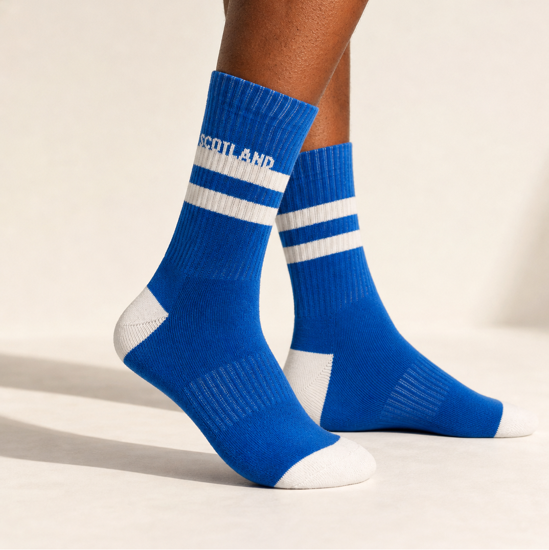 A pair of bright blue crew socks with white stripes and white toe/heel sections, worn on a model's legs against a clean off-white studio background. The word 'SCOTLAND' is printed in white on the front sock.