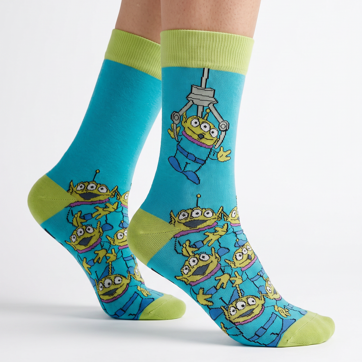 A pair of crew socks worn on a person's feet, featuring a bright blue background with a repeating pattern of cartoon green alien characters and lime green cuffs, heels, and toes on a light gray studio background.