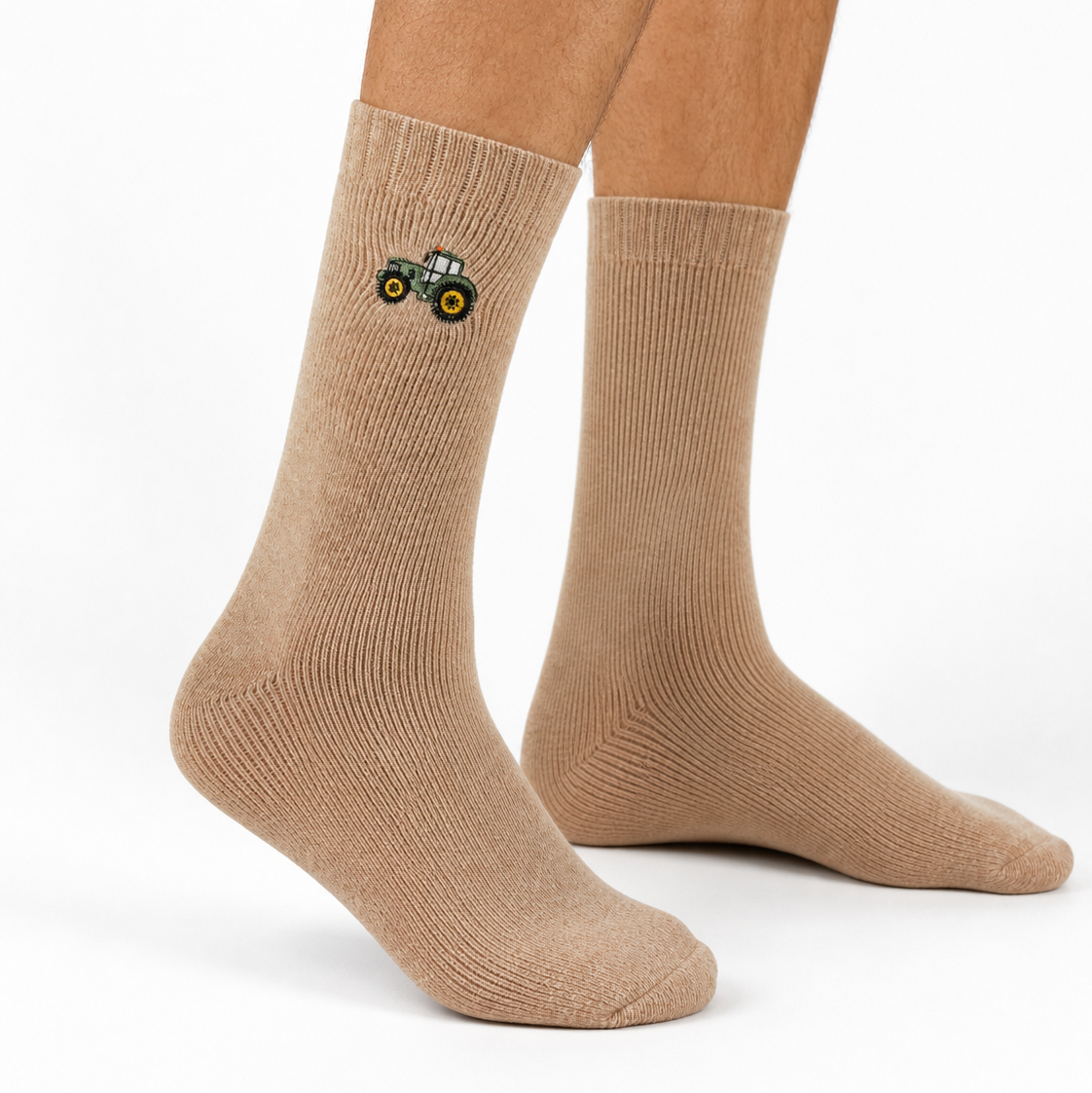 A pair of beige crew socks worn on a person's legs, featuring a small embroidered green tractor graphic on the left sock.