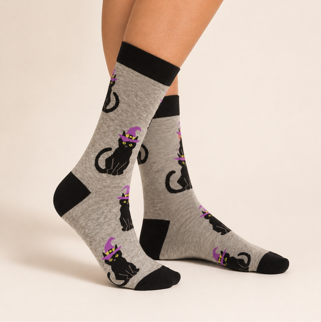 A pair of crew socks worn on a model's feet, featuring a gray heather base with black cat illustrations wearing purple witch hats, set against a plain beige studio background.